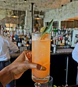 The hand of a Black woman holding an orange cocktail in a glass in front of a modern bar in an Italian restaurant on a weekend getaway to Buffalo, New York.